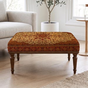 Puede incluir: Un otomán rectangular con una tapicería de tonos cálidos y ricos que presenta un intrincado patrón de medallón floral simétrico en tonos naranja intenso, dorado y marrón, apoyado sobre cuatro patas de madera torneada.