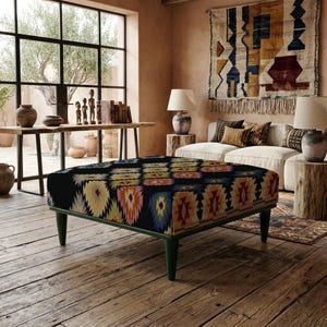 Puede incluir: Un otomán cuadrado con base de tela negra y un patrón geométrico repetitivo en amarillo, naranja, rojo, azul y blanco. La pieza se apoya sobre cuatro patas cónicas de color verde oscuro, proporcionando una base estable para el asiento.