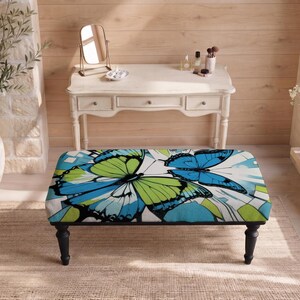 May include: A rectangular ottoman featuring a vibrant graphic print of large blue and lime green butterflies on a white background, supported by four black turned wooden legs. The upholstery displays a bold, colorful pattern of wings.