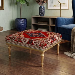 Puede incluir: Un otomán cuadrado con una superficie textil de colores rojo vibrante, azul marino y crema, con un diseño inspirado en alfombras tradicionales. La pieza se apoya sobre cuatro patas cónicas de metal dorado que ofrecen una base estable.