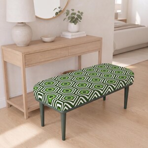 May include: A rectangular bench featuring a cushioned seat with a repeating geometric pattern of green, dark green, and off-white hexagons. The bench is supported by four slender, dark grey tapered legs that provide a stable base for the seat.