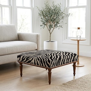 Op de afbeelding: Een vierkante poef met een zwart-witte zebraprint stoffering. Het meubelstuk wordt ondersteund door vier donkerbruine houten poten met een gedraaid, taps toelopend ontwerp. Het bovenoppervlak is vlak en bekleed met de textielstof.