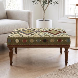 Puede incluir: Un otomán rectangular con una parte superior de color crema con un patrón geométrico de diamantes en tonos rojo, azul y bronceado. Los lados son de color verde oliva con un motivo de diamantes repetido, sobre cuatro patas de madera torneada.