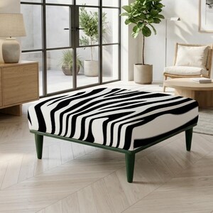 Puede incluir: Un otomano cuadrado con un llamativo patrón de rayas de cebra en blanco y negro en la parte superior tapizada. La pieza se apoya sobre cuatro patas cónicas de color verde oscuro que proporcionan una base estable para el asiento bajo.