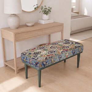May include: A rectangular bench featuring a cushioned seat upholstered in a multi-coloured fabric with an intricate floral and geometric pattern. The bench is supported by four tapered, dark green wooden legs that provide a stable base.