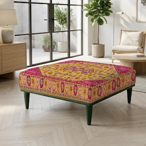 Op de afbeelding: Een vierkante poef met een levendige gele en roze stoffen bekleding voorzien van geometrische en florale patronen. Het meubelstuk rust op vier donkergroene, taps toelopende poten die een lage basis vormen voor dit gestoffeerde item.