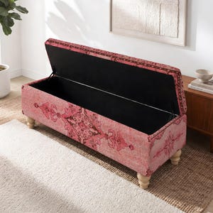Puede incluir: Un banco de almacenamiento rectangular tapizado en tela rosa con un intrincado patrón rojo oscuro. El banco cuenta con una tapa abatible que revela un compartimento interior negro y se apoya sobre cuatro patas de madera clara.