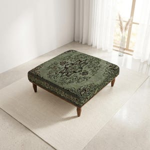 Puede incluir: Un otomán cuadrado con tapicería de tela verde salvia y un intrincado patrón floral y de medallones en color marrón oscuro. La pieza se apoya sobre cuatro patas de madera cortas y cónicas con un acabado marrón medio, de perfil bajo.