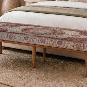 May include: A rectangular bench featuring a cushioned seat upholstered in a fabric with a detailed red, cream, and grey ornate pattern. The bench is supported by four light brown wooden legs that taper slightly toward the floor.