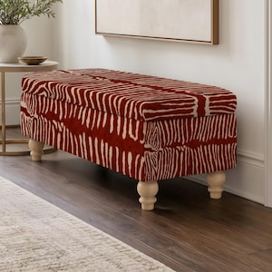 May include: A rectangular storage bench upholstered in a deep red fabric with a white zebra stripe pattern. The bench features a hinged lid and is supported by four light-colored, rounded wooden bun feet that elevate the base from the floor.