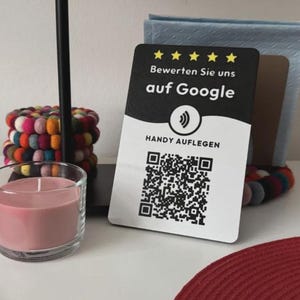 Könnte beinhalten: Ein rechteckiges schwarz-weißes Schild mit fünf gelben Sternen, dem Text "Bewerten Sie uns auf Google", einem Kontaktlos-Symbol, dem Text "HANDY AUFLEGEN" und einem QR-Code. Das Schild steht neben einer rosa Kerze in einem Glas.