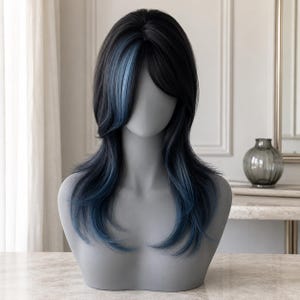 May include: A shoulder-length wig featuring layered black hair with prominent blue highlights framing the face and blue tips. The hair is styled with a side-swept fringe and soft waves that cascade down the sides, displayed on a gray mannequin head.