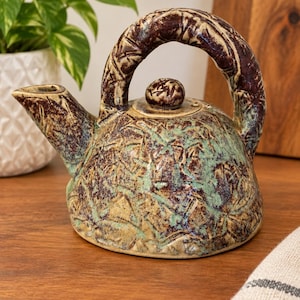 May include: A ceramic teapot featuring a textured, mottled glaze in shades of sage green, tan, and dark brown. The teapot has a rounded body, a curved spout, a thick arched handle, and a small circular lid with a central knob.
