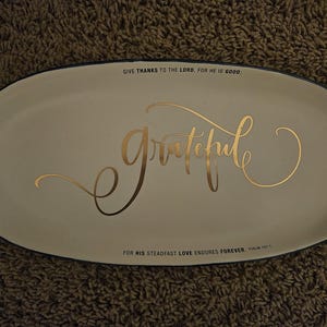 Könnte beinhalten: Ein ovales, cremefarbenes Keramiktablett mit dunklem Rand. In der Mitte steht das Wort grateful in goldener Schreibschrift. Der obere Text lautet GIVE THANKS TO THE LORD, FOR HE IS GOOD, und der untere Text lautet FOR HIS STEADFAST LOVE ENDURES FOREVER. PSALM 107:1.