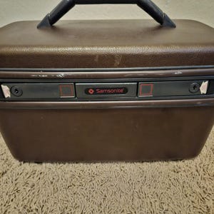 May include: A brown hardshell vanity case with a dark brown top handle. The front features a central dark grey locking mechanism with the red Samsonite logo, flanked by two sliding latches. The exterior has a textured finish with visible wear.