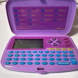 May include: A purple electronic diary with a flip-top lid featuring a menu of functions including Daily Diary, Calculator, and Horoscope. The base includes a QWERTY keyboard with blue circular keys, a digital screen, and purple directional buttons.