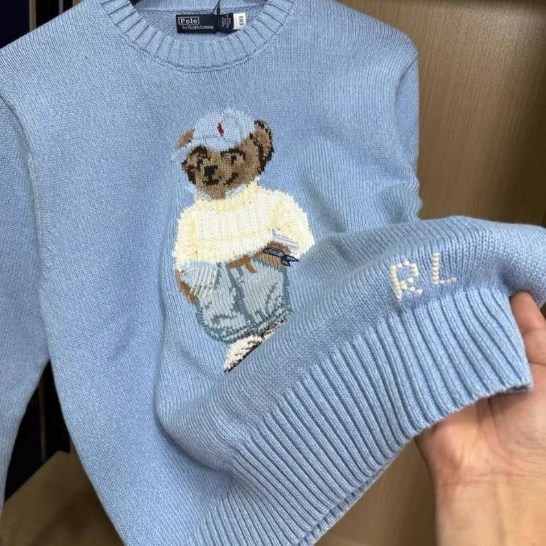 Puede incluir: Un su&eacute;ter de punto de manga larga en color azul claro con cuello redondo y borde acanalado. El frente muestra un oso de peluche tejido en intarsia con gorra, su&eacute;ter crema y jeans azules. Las letras RL est&aacute;n bordadas en blanco en el pu&ntilde;o acanalado.