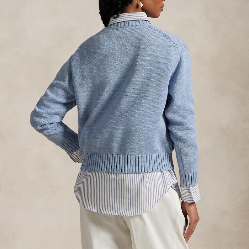Puede incluir: Un su&eacute;ter de punto de manga larga en color azul claro con cuello redondo, pu&ntilde;os y dobladillo acanalados, superpuesto sobre una camisa de botones con rayas verticales blancas y grises y dobladillo curvo, con pantalones blancos, visto de espaldas.