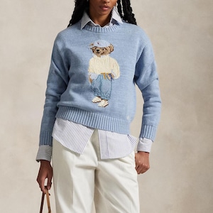 Puede incluir: Un su&eacute;ter de punto de manga larga en color azul claro con un gr&aacute;fico central de un oso de peluche que viste un su&eacute;ter de punto trenzado color crema, jeans azules y una gorra azul claro. El su&eacute;ter tiene cuello redondo, pu&ntilde;os y dobladillo acanalados.