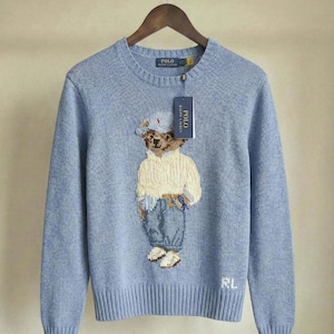 Puede incluir: Un su&eacute;ter de manga larga y cuello redondo en color azul claro con un gr&aacute;fico tejido de un oso de peluche que viste un su&eacute;ter color crema, pantalones azules y una gorra. El su&eacute;ter tiene una etiqueta Polo Ralph Lauren y las letras RL bordadas.