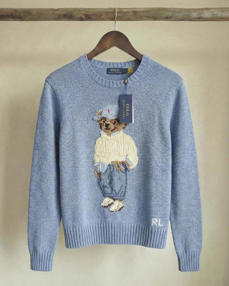 Puede incluir: Un su&eacute;ter de manga larga y cuello redondo en color azul claro con un gr&aacute;fico tejido de un oso de peluche que viste un su&eacute;ter color crema, pantalones azules y una gorra. El su&eacute;ter tiene una etiqueta Polo Ralph Lauren y las letras RL bordadas.