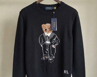 Puede incluir: Un suéter negro de manga larga con cuello redondo que presenta un gráfico tejido de un oso de peluche con traje. El suéter tiene cuello, puños y dobladillo acanalados, con las letras RL bordadas en blanco en la parte inferior y una etiqueta Polo Ralph Lauren.