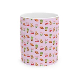 Puede incluir: Una taza cilíndrica de cerámica con fondo rosa claro que presenta un patrón repetitivo de acuarela con ositos de peluche marrones, cerezas rojas con lazos rosas, lattes de matcha verdes, tulipanes rosas y croissants dorados en el exterior.