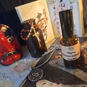 May include: A small amber glass spray bottle with a beige label reading Bayrum Protection renforcée énergétique, positioned next to a silver pendant, a red and black mask, a brown glass jar with beads, and a gold wax seal stamp.