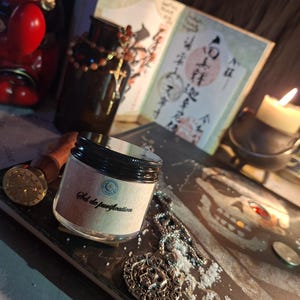 May include: A small clear glass jar with a black lid and a beige label featuring the text Sel de purification. The jar is surrounded by a silver skull pendant on a chain, loose salt grains, a wax seal stamp, a dark bottle, and a lit candle.