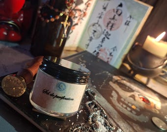 May include: A small clear glass jar with a black lid and a beige label featuring the text Sel de purification. The jar is surrounded by a silver skull pendant on a chain, loose salt grains, a wax seal stamp, a dark bottle, and a lit candle.