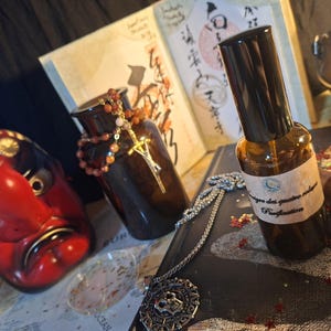 May include: A brown glass spray bottle with a label reading "Purification" sits next to a red Tengu mask, a dark glass jar with a beaded necklace, a silver pendant, and an open book with Japanese calligraphy and a small metal cauldron.