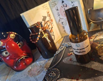 Puede incluir: Un frasco de spray de vidrio marrón con una etiqueta que dice "Purification" junto a una máscara Tengu roja, un frasco de vidrio oscuro con un collar de cuentas, un colgante de plata, un libro con caligrafía y un caldero pequeño.