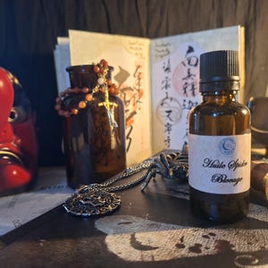 May include: A small amber glass bottle with a black cap and a white label reading Huile Spider Blocage. The bottle sits near a red tengu mask, a brown glass jar with a beaded rosary, a silver pendant necklace, and a metal wax seal stamp.