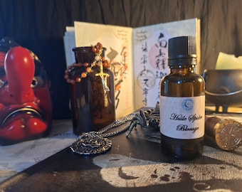 Puede incluir: Un frasco pequeño de vidrio ámbar con tapa negra y etiqueta blanca que dice Huile Spider Blocage. El frasco se encuentra junto a una máscara tengu roja, un frasco de vidrio marrón con un rosario, un collar de plata y un sello de metal.