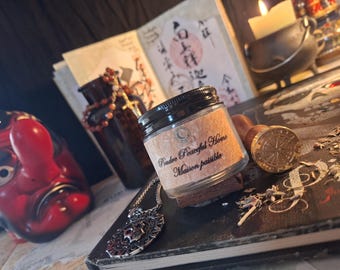 Puede incluir: Un pequeño frasco de vidrio con tapa negra y etiqueta beige que muestra el texto Poudre Peaceful Home Maison paisible. El frasco está colocado cerca de un sello de lacre dorado, un colgante plateado y una máscara roja de nariz larga.