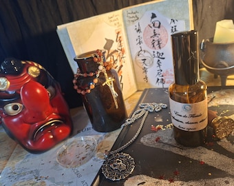 May include: A brown glass spray bottle with a label reading Eau de Floride, a red Tengu mask, a dark glass vase with a beaded cross necklace, a silver skull pendant necklace, a wax seal stamp, and an open book with Asian calligraphy.
