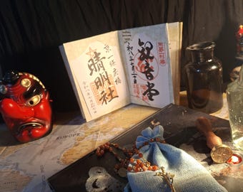 Puede incluir: Una máscara tengu roja, un libro abierto con caligrafía japonesa, una bolsa de tela azul claro que contiene un collar de cuentas con un colgante de cruz dorada, un sello de cera y una botella de vidrio oscuro sobre un mapa del mundo.