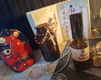 May include: A brown glass spray bottle with a label reading Fast luck Chance rapide, positioned next to a red and black Tengu mask, a dark glass jar draped with a beaded necklace featuring a gold-tone sword pendant, and a wax seal stamp.