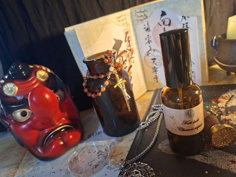 May include: A brown glass spray bottle with a label reading Fast luck Chance rapide, positioned next to a red and black Tengu mask, a dark glass jar draped with a beaded necklace featuring a gold-tone sword pendant, and a wax seal stamp.