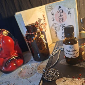 Könnte beinhalten: Eine rot-schwarze Tengu-Maske, eine braune Glasflasche mit Perlenkette, eine kleine bernsteinfarbene Tropfflasche mit dem Etikett Huile Allipie, ein silberner Totenkopfanhänger an einer Kette und ein goldener Wachssiegelstempel auf einer Karte.