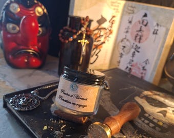 Puede incluir: Un pequeño frasco de vidrio con tapa negra y una etiqueta que dice Poudre d'Hermès Protection en voyage, acompañado de un sello de cera de madera, un colgante de calavera plateado, una máscara roja y negra y una botella oscura sobre un libro.