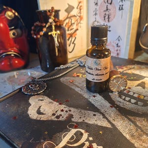May include: A small amber glass bottle with a black cap and a beige label reading Huile Van-Van, positioned on a black book cover featuring a white skull and crossbones graphic, alongside a metal wax seal stamp and a silver chain necklace.