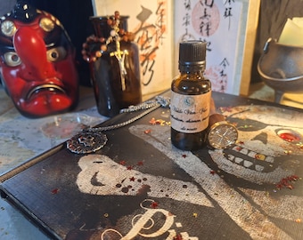 Puede incluir: Un pequeño frasco de vidrio ámbar con tapa negra y etiqueta beige que dice Huile Van-Van, colocado sobre una cubierta de libro negra con un gráfico de calavera blanca, junto a un sello de metal y un collar de cadena de plata.