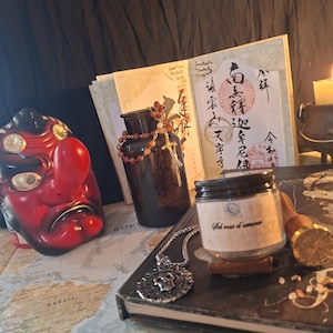Könnte beinhalten: Eine rot-schwarze Tengu-Maske, eine dunkelbernsteinfarbene Glasflasche mit Perlenkette, ein kleines Glas mit der Aufschrift Sel rose d'amour, ein silberner Totenkopfanhänger, ein Wachssiegelstempel und ein offenes Buch mit japanischer Kalligrafie.