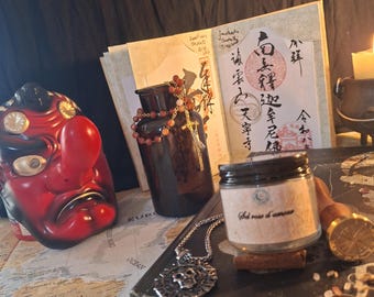 Puede incluir: Una máscara Tengu roja y negra, una botella de vidrio ámbar oscuro con un collar de cuentas, un frasco pequeño con la etiqueta Sel rose d'amour, un colgante de calavera plateado, un sello de cera y un libro abierto con caligrafía japonesa.