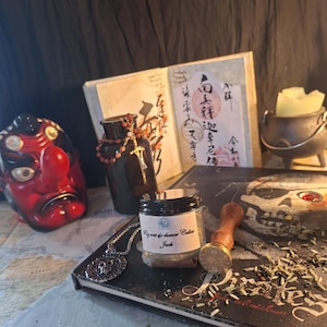 May include: A small glass jar with a label reading "Pig vert de chance Calico Jack" sits on a book titled "Pirates" next to a metal wax seal stamp, a silver skull pendant, a dark bottle with a beaded cross, a red tengu mask, and a small cauldron.