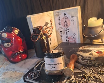 May include: A small glass jar with a label reading "Pig vert de chance Calico Jack" sits on a book titled "Pirates" next to a metal wax seal stamp, a silver skull pendant, a dark bottle with a beaded cross, a red tengu mask, and a small cauldron.