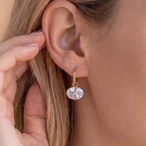 May include: A gold hoop earring featuring a dangling oval-shaped clear gemstone. The earring is secured through the earlobe, showcasing a polished metallic finish and a faceted stone that reflects light, creating a bright and clear appearance.