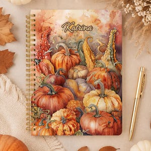 Peut inclure: Un carnet à spirales orné d'une illustration à l'aquarelle représentant diverses citrouilles et courges dans des tons orange, jaune et vert. Le prénom Katrina est imprimé dans une police cursive sombre en haut au centre. Un stylo doré est posé à côté.