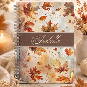 Peut inclure: Un carnet à reliure spirale orné d'un motif aquarelle de feuilles d'érable automnales dans des tons orange, rouge et jaune. Une bande horizontale brune au centre affiche le prénom Isabella en écriture cursive blanche.